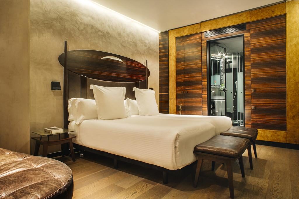 Quarto do Hotel Bagués, com cama de casal, mesas de cabeceira de ambos os lados, bancos de couro aos pés e um roupeiro de madeira ao lado.