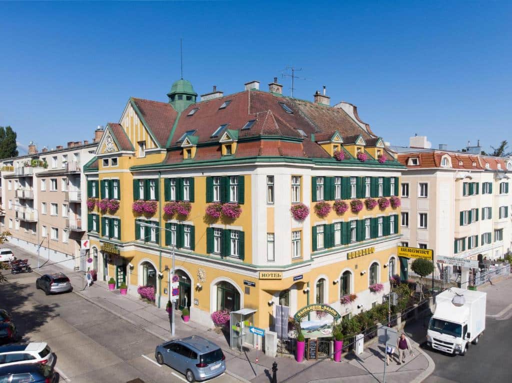 Edifício amarelo do Hotel Bergwirt Schönbrunn, com persianas verdes e floreiras, numa esquina, num dia soalheiro.