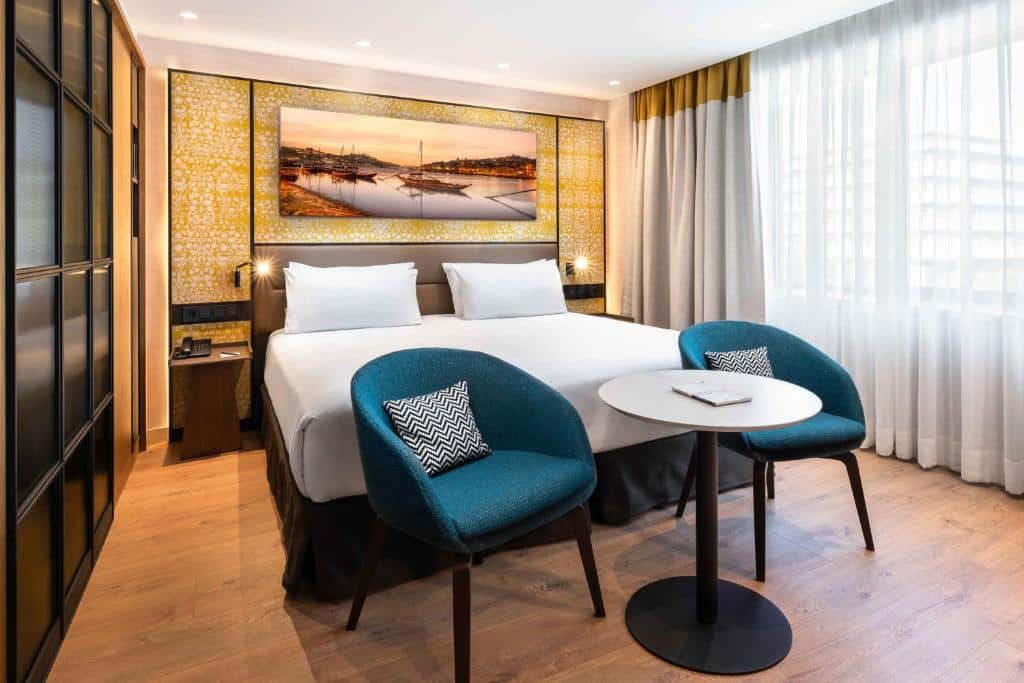 Quarto do Eurostars Aliados com cama de casal e duas cadeiras azuis junto a uma mesa redonda aos pés da cama.