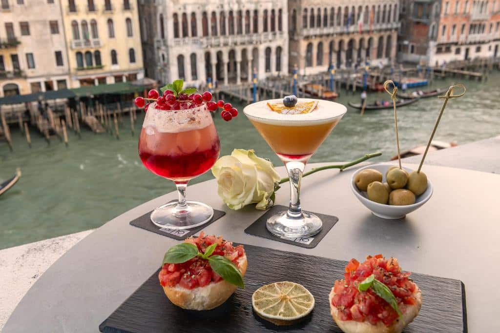 Duas bebidas coloridas em copos, uma vermelha com frutos vermelhos e outra amarela com uma rodela de limão, numa mesa de madeira com azeitonas e bruschetta, com o Grande Canal de Veneza ao fundo