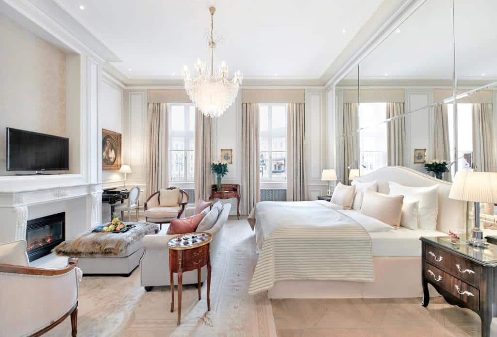 Quarto elegante do Hotel Sacher Wien, com cama, sofá, lustre, lareira, TV, janelas grandes e uma parede espelhada.