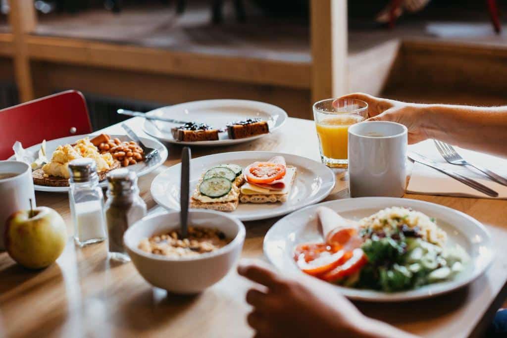 Mesa posta com opções de pequeno-almoço no ClinkNOORD Hostel, com torradas, fruta, saladas, cereais, café e uma mão a segurar um copo de sumo de laranja.