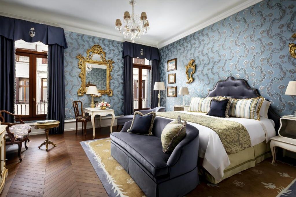 Quarto luxuoso com cama de casal, paredes com padrão azulado, espelho dourado e decoração elegante