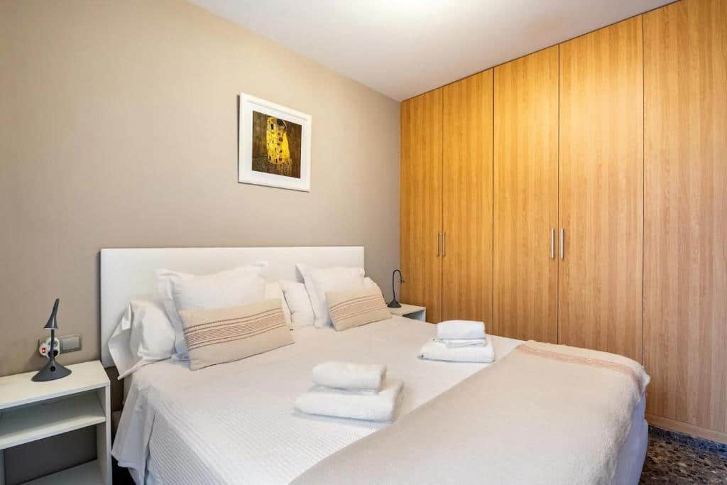Quarto do Moderno apartamento con vistas al mar, com paredes bege, armário de madeira e cama de casal branca com almofadas e colcha às riscas.