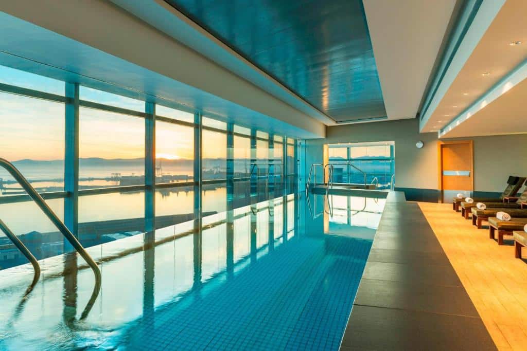Piscina interior no The Westin Cape Town com espreguiçadeiras e grandes janelas com vista para a marginal ao pôr do sol.