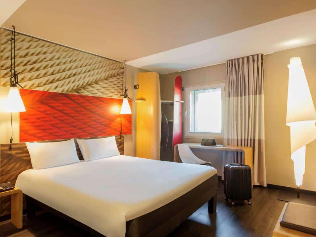 Quarto do Hotel Ibis Lisboa Liberdade com cama de casal, dois candeeiros, pequena zona de trabalho e janela, com detalhes em vermelho e bege.