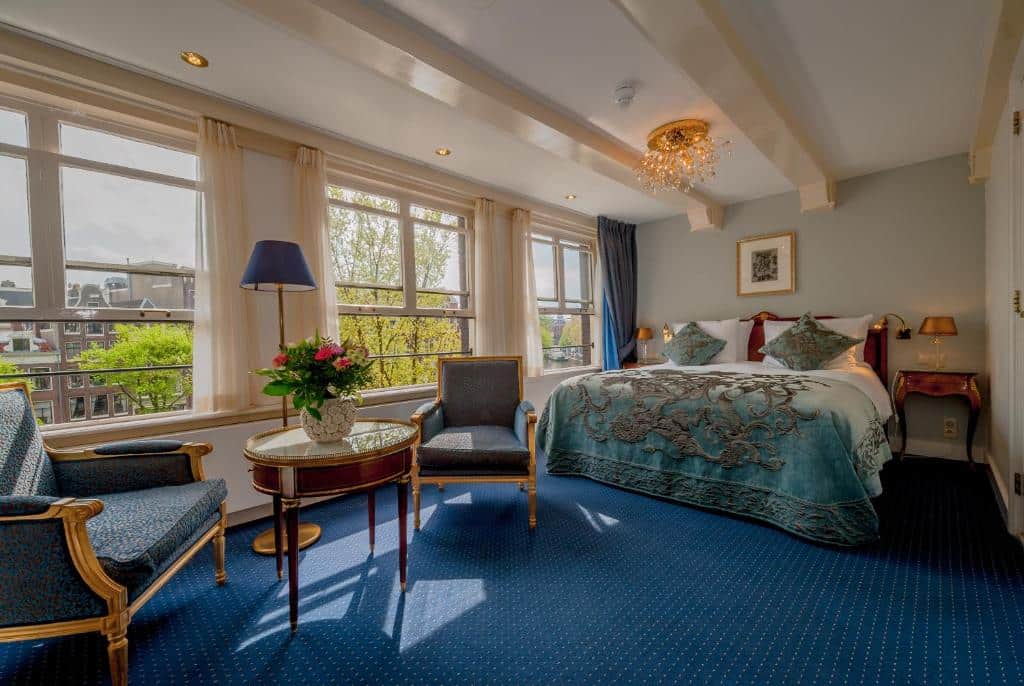 Quarto Duplo Deluxe Superior do Ambassade, de 25 m², com cama de casal grande, dois cadeirões de época, mesa redonda e janela com vista para árvores e edifícios históricos