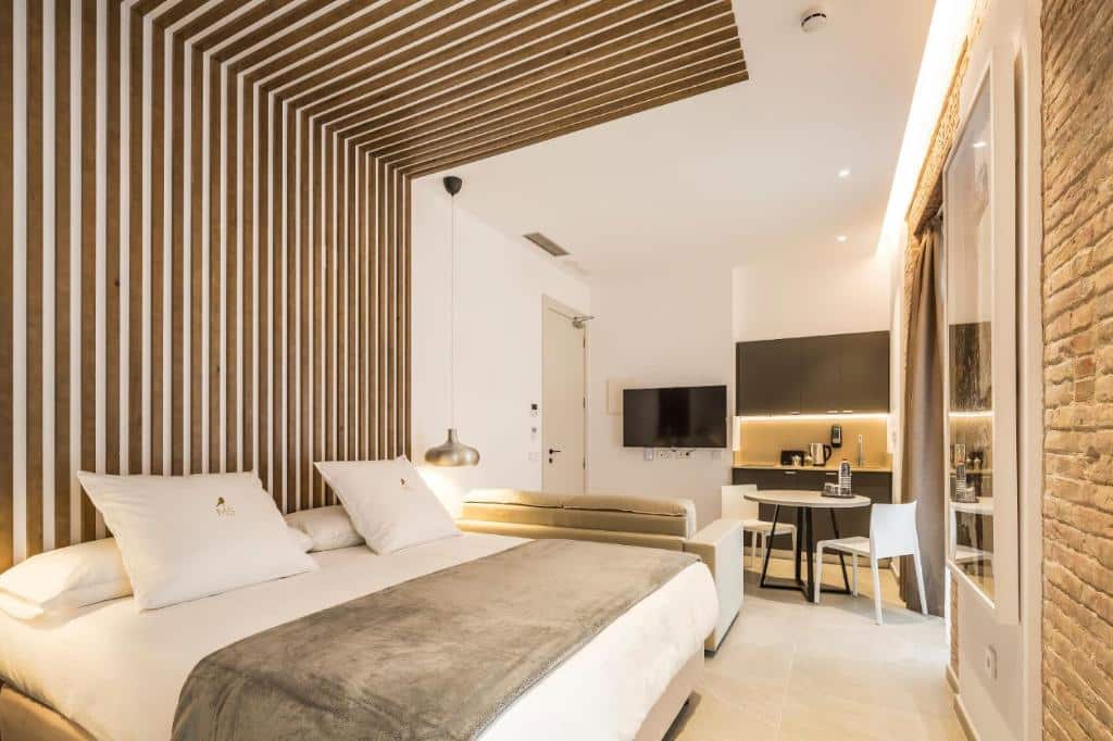 Quarto moderno do Mon Suites Benlliure, com paredes de madeira clara, cama de casal e móveis em tons neutros, uma mesa com duas cadeiras brancas e televisão na parede.