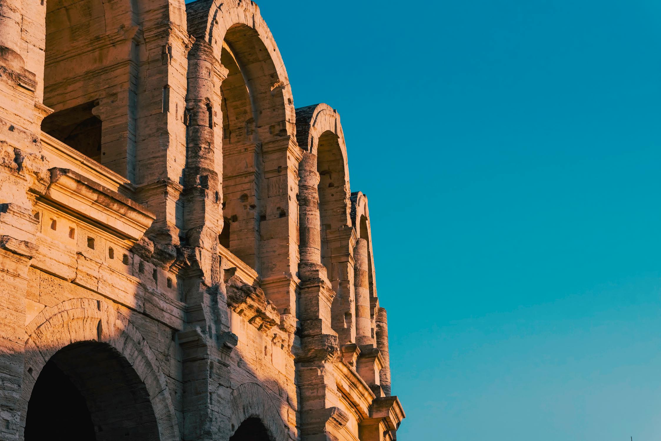 Exterior de um antigo anfiteatro romano com arcos de pedra, iluminado pela luz quente do sol, em Arles.
