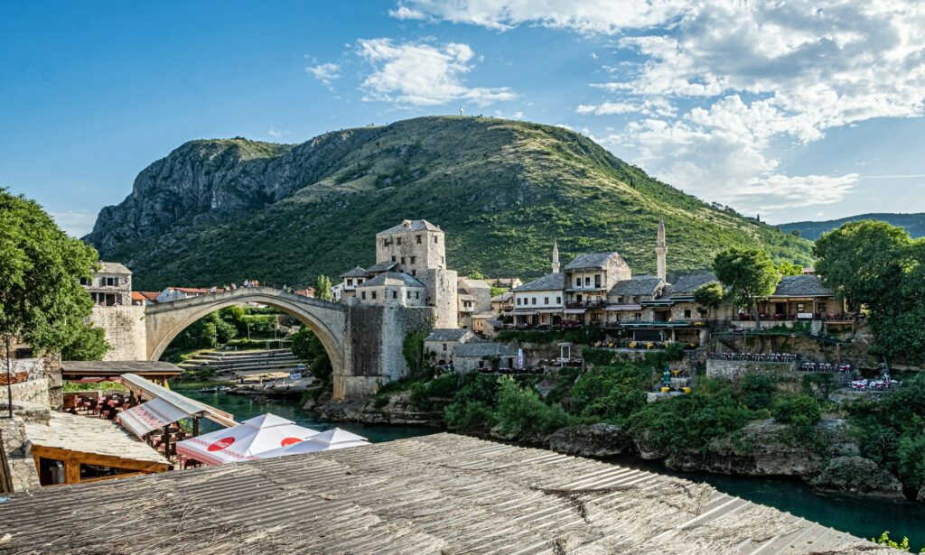 Panorama da cidade histórica de Mostar, na Bósnia e Herzegovina, destacando a famosa Stari Most, ponte em arco de pedra que atravessa o rio Neretva. À volta da ponte, há edifícios de pedra com telhados de telha, incluindo uma mesquita com um minarete visível. O cenário é emoldurado por montanhas verdes.