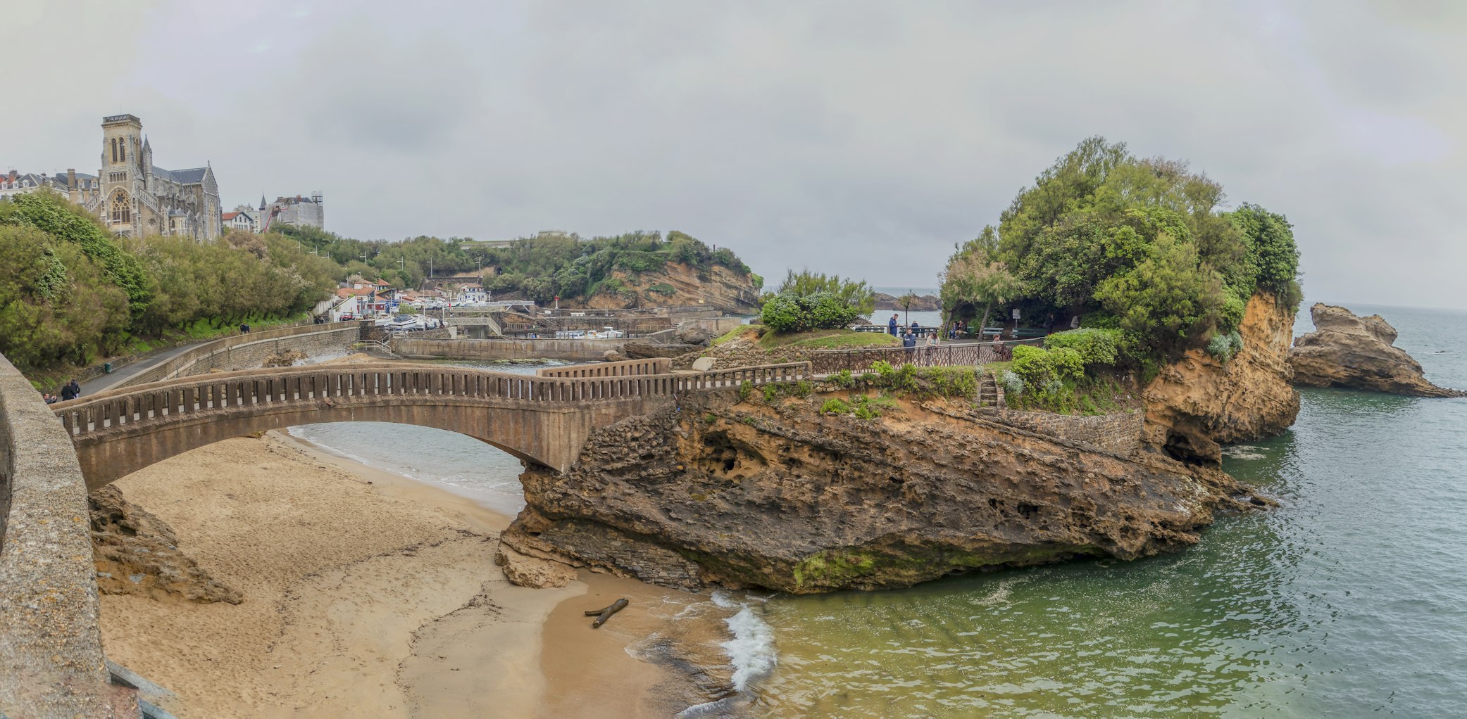 Ponte de pedra a ligar uma praia de areia a um promontório rochoso com vegetação, em Biarritz.