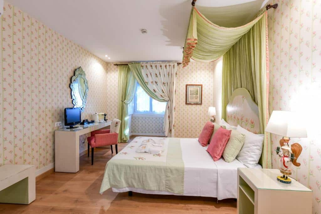 Quarto do Casa de São Mamede Hotel com uma cama de casal com dossel em tecido verde-claro, uma janela com cortinas no mesmo tom, uma bancada com televisão e espelho e uma poltrona; o chão imita madeira.