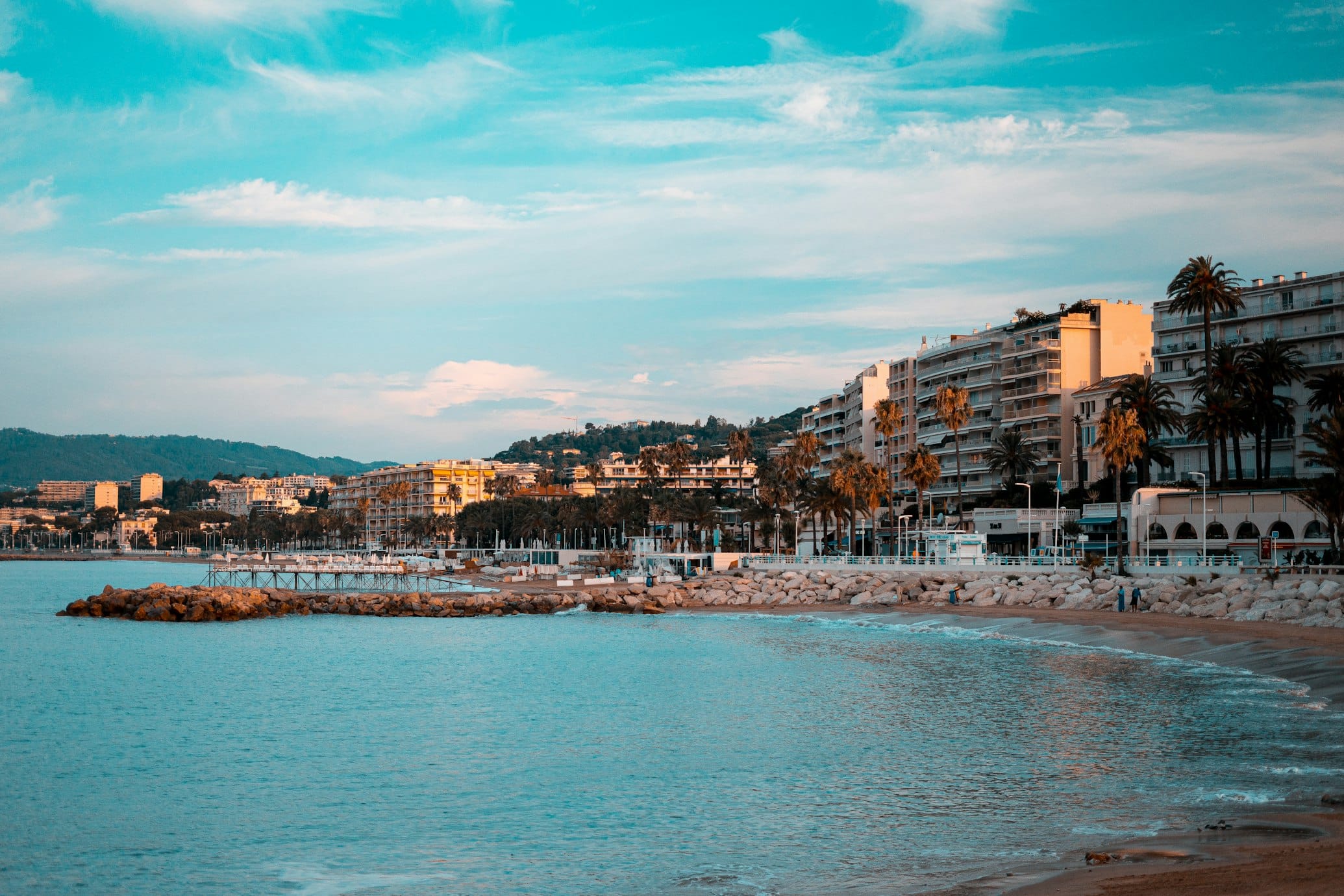 Paisagem costeira de Cannes, com praia de areia, palmeiras e edifícios alinhados ao longo da costa.