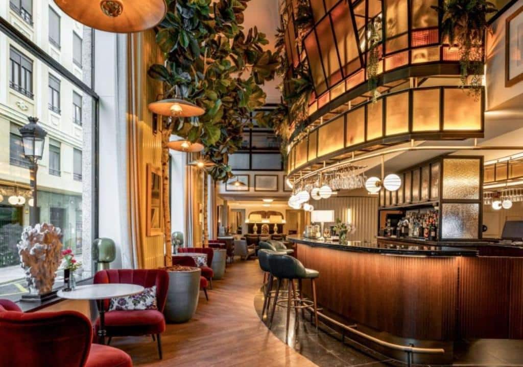 Saguão luxuoso do Only YOU Hotel Valencia, de decoração clássica, com sofás de veludo vermelho, móveis de madeira, plantas suspensas e um amplo bar de balcão de madeira com bancos e vários lustres circulares.