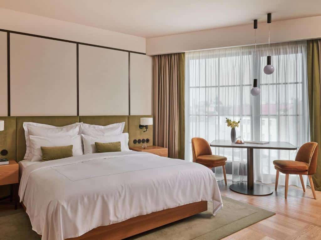Quarto de hotel moderno e espaçoso do The Julius Prague, com cama de casal, mesa com duas cadeiras e uma janela com cortinas verdes.