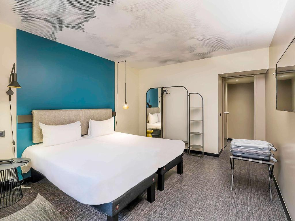 Quarto do Ibis Lisboa Saldanha com uma cama de casal, um espelho de corpo inteiro junto a um bengaleiro, mesas de cabeceira com candeeiros e uma televisão
