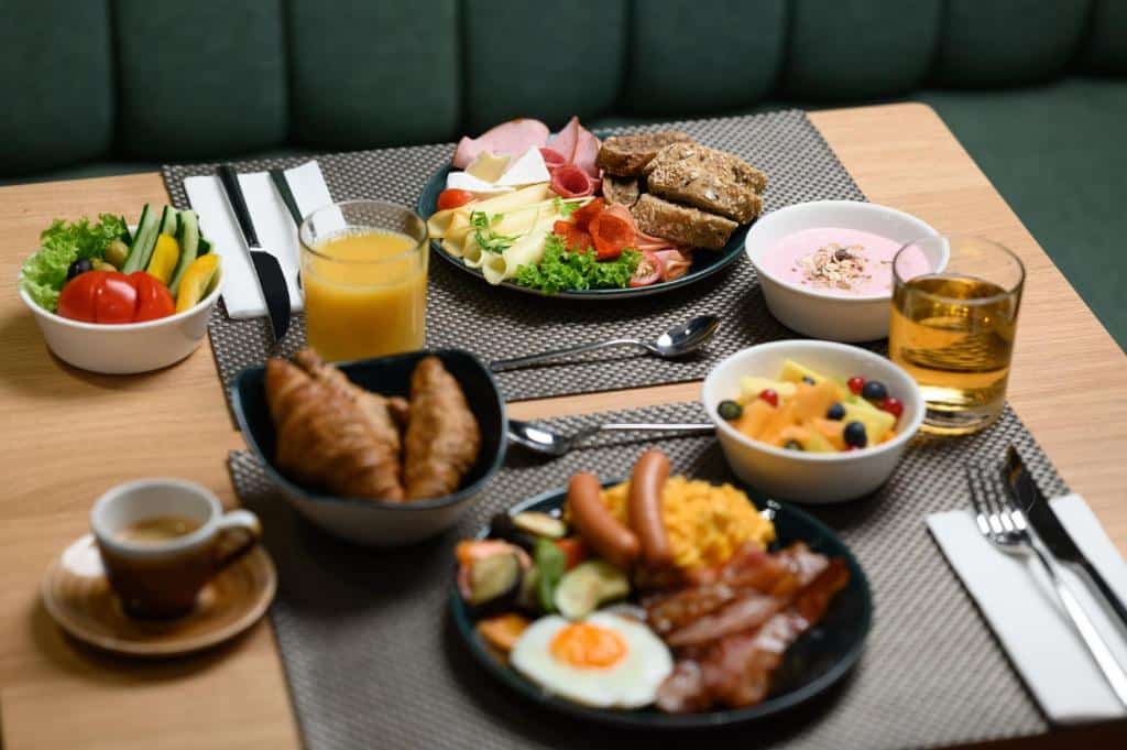 Mesa do Hotel Oktogon Haggenmacher posta para duas pessoas, com um pequeno-almoço que inclui croissants, ovos, fruta, charcutaria, queijo, sumo e café.