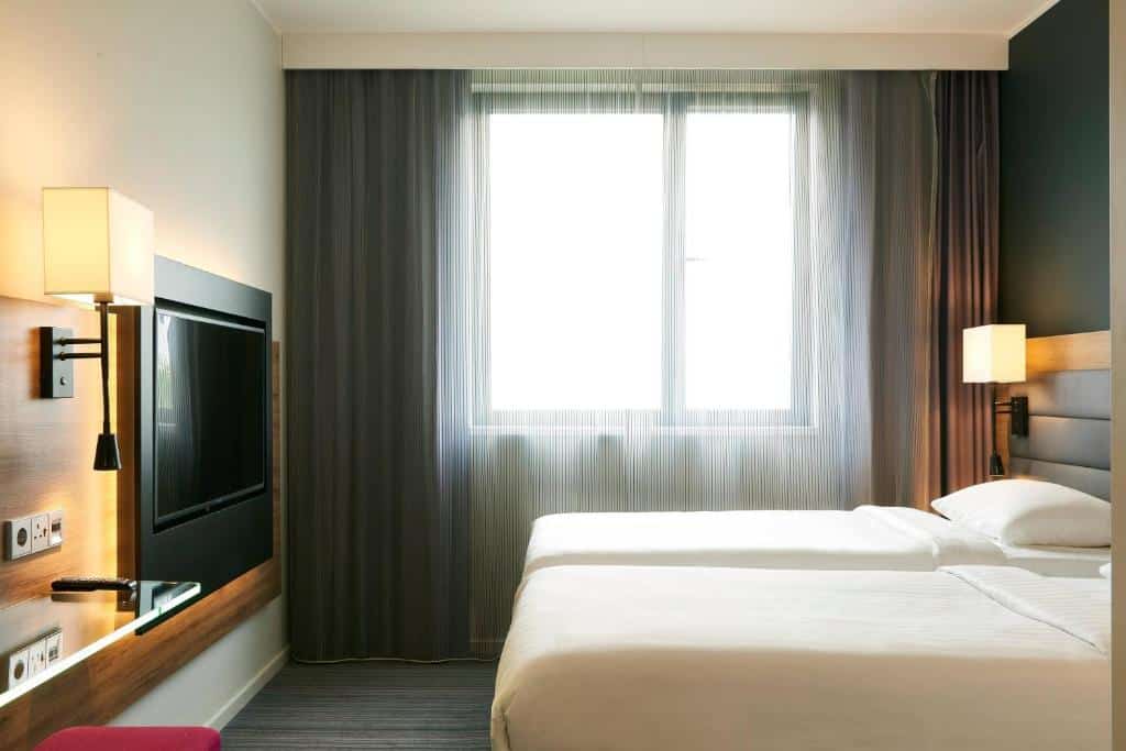 Quarto do Moxy Milan Linate Airport com duas camas de solteiro, janela ampla com cortinas e televisão em frente às camas