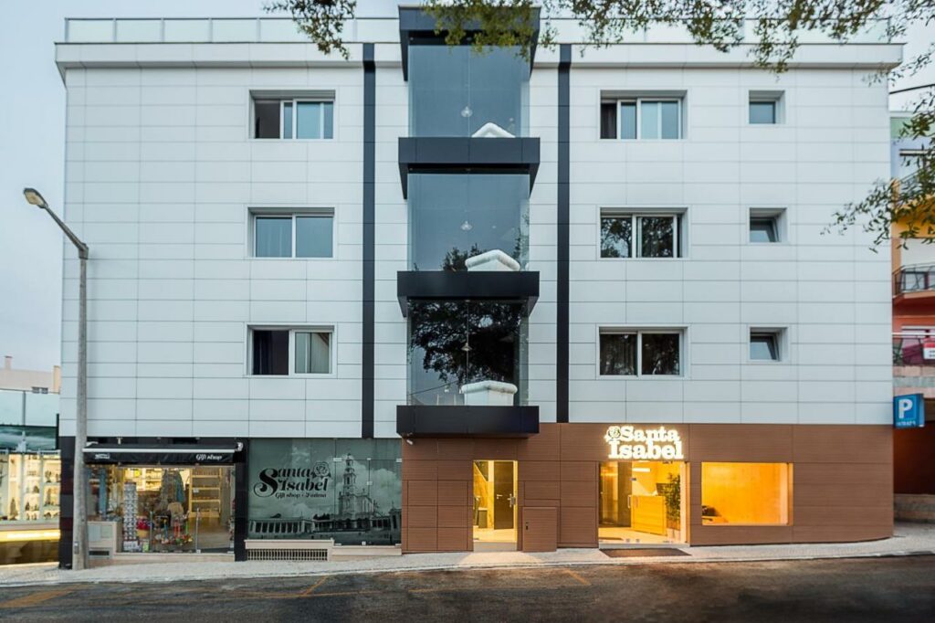 Edifício moderno de quatro pisos com escadaria central em vidro, lojas no rés do chão e um letreiro iluminado com o nome Santa Isabel.