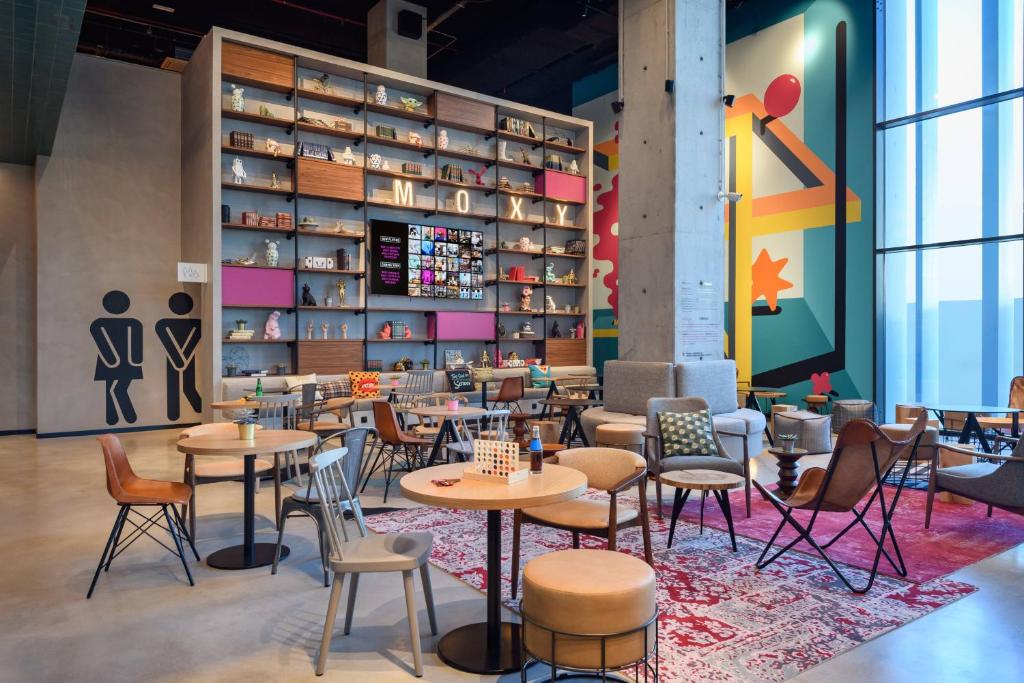 Zona comum do Moxy Lisboa Oriente com vários sofás, poltronas, pequenas mesas redondas com cadeiras, uma estante ampla com elementos decorativos e uma janela do chão ao teto do lado direito.
