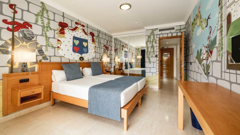 Quarto do Ukino Palmeiras Village – Family Resort, com cama de casal do lado esquerdo da imagem com uma cômoda de madeira de cada lado da cama. Representa hotéis all inclusive em Portugal.