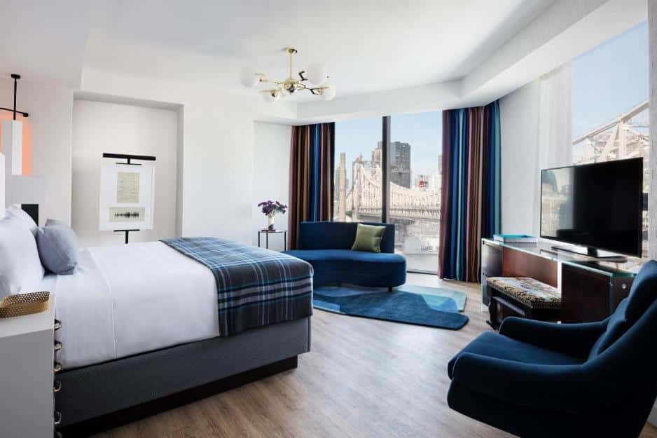 Na suíte do Graduate New York há uma cama de casal ao centro, com cabeceira estofada e roupa branca. À direita, uma poltrona azul e uma TV sobre um suporte; à esquerda, uma mesa de cabeceira com candeeiro. A janela ao fundo tem vista para a cidade.
