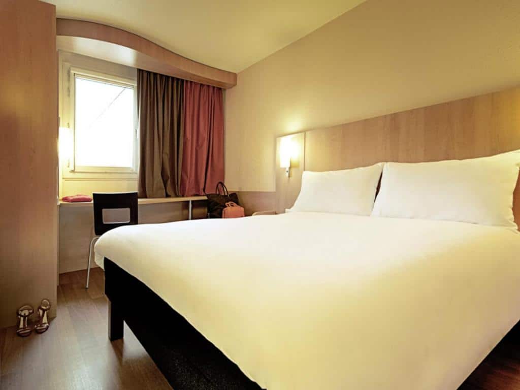 Quarto do ibis Porto Centro São Bento com cama de casal do lado direito e do lado esquerdo da cama uma mesa de trabalho. Representa hotéis ibis no Porto.