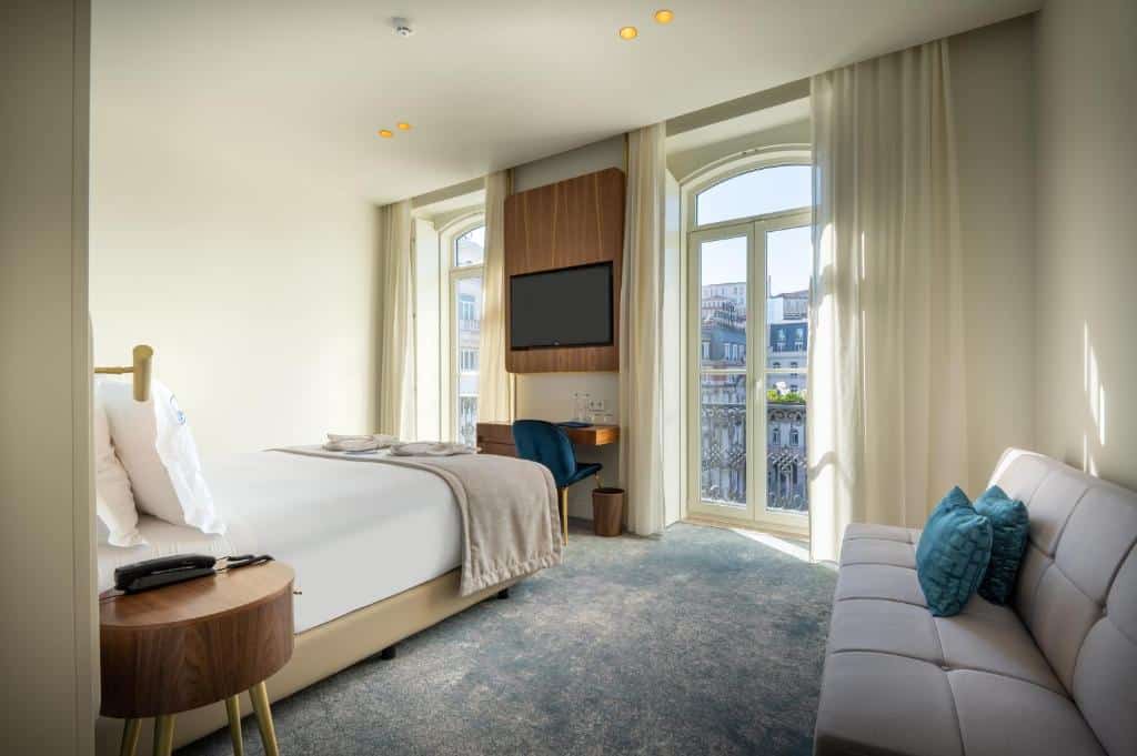 Quarto do Blue Liberdade Hotel com duas varandas e, entre elas, uma televisão e uma secretária com cadeira; ao centro, uma cama de casal e, à direita, um pequeno sofá com duas almofadas