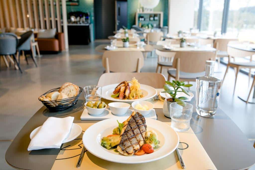 Uma mesa para duas pessoas com peixe grelhado, pão, azeitonas, sobremesa e água numa zona de refeições moderna e iluminada de um restaurante no Mercure Fátima.