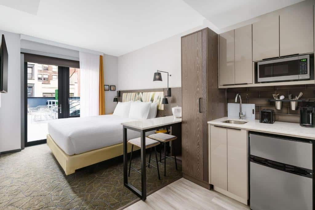 No quarto do TownePlace Suites by Marriott New York Manhattan Chelsea há uma cama de casal com cabeceira estofada ao centro. À esquerda, uma pequena cozinha com armários, micro-ondas e lava-loiça; à direita, uma secretária com cadeira preta. Ao fundo, uma janela grande com cortinados.