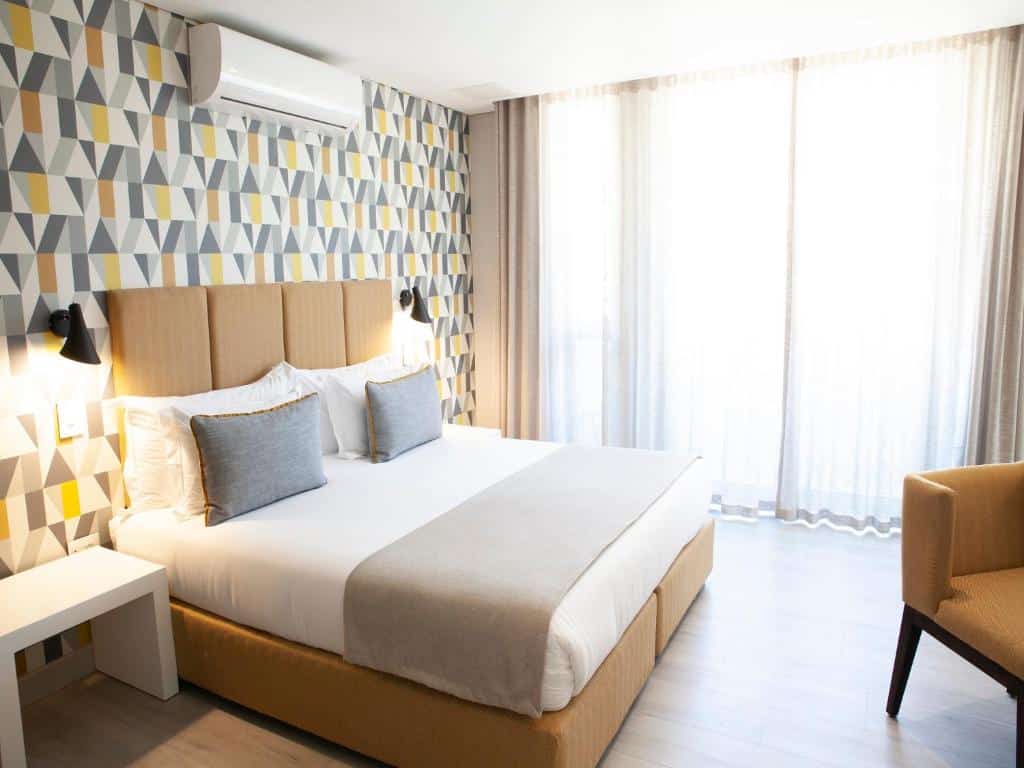 Quarto do Zest Boutique Hotel com cama de casal, parede com detalhes geométricos, dois candeeiros, janela com cortinas e uma poltrona.