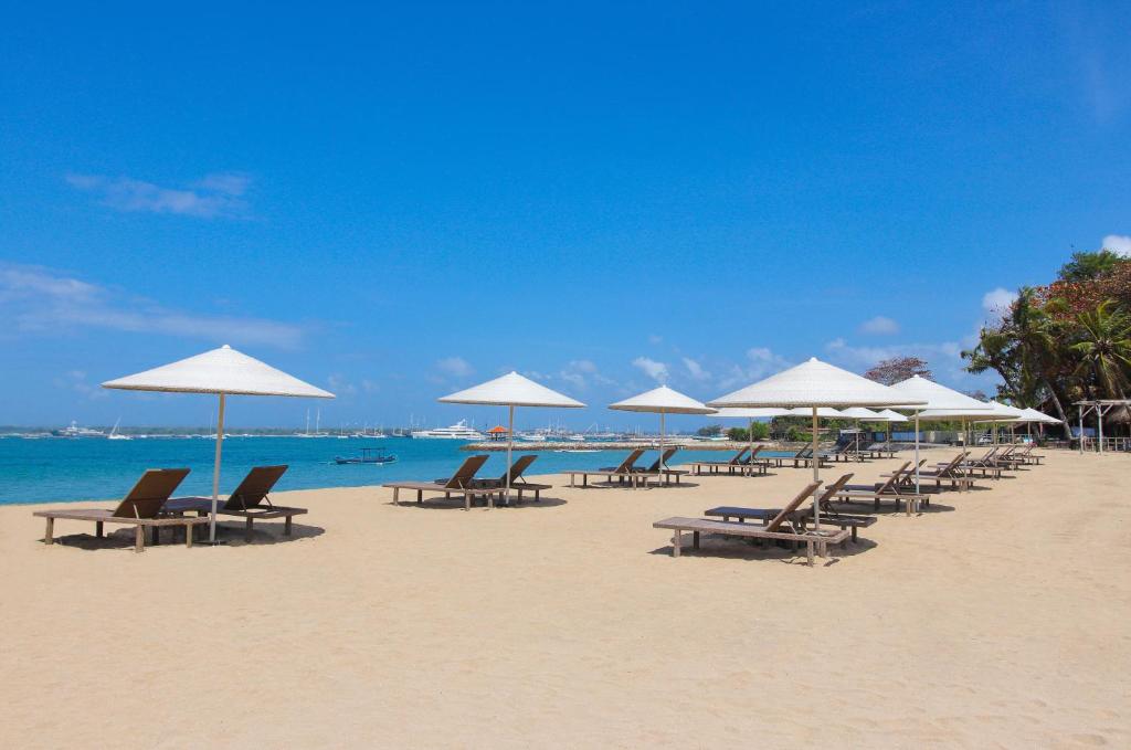 Espreguiçadeiras sob guarda-sóis brancos numa praia de areia em Sanur, com céu azul e mar calmo, em Bali.