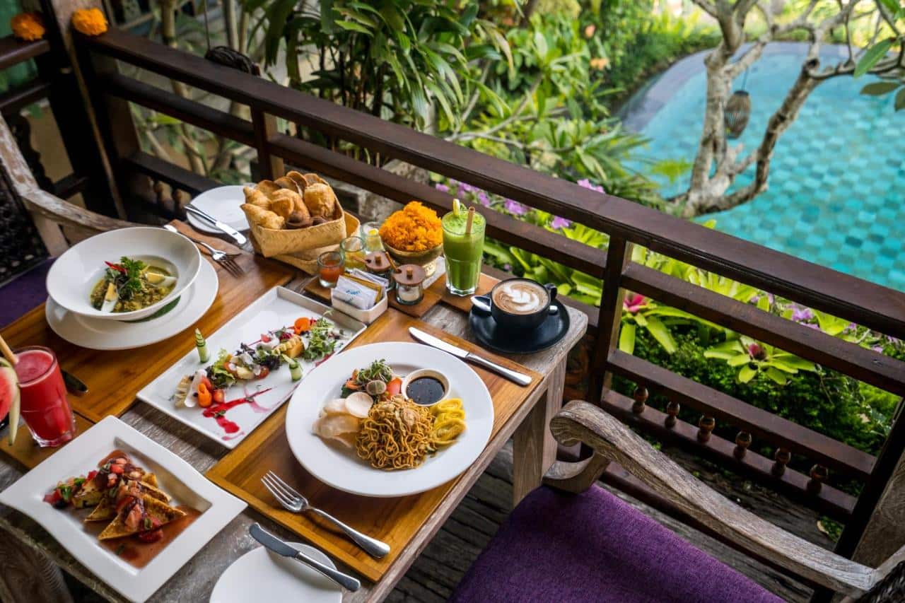 Mesa de madeira com pratos e bebidas e vista para uma piscina rodeada de vegetação, num hotel em Ubud, Bali.