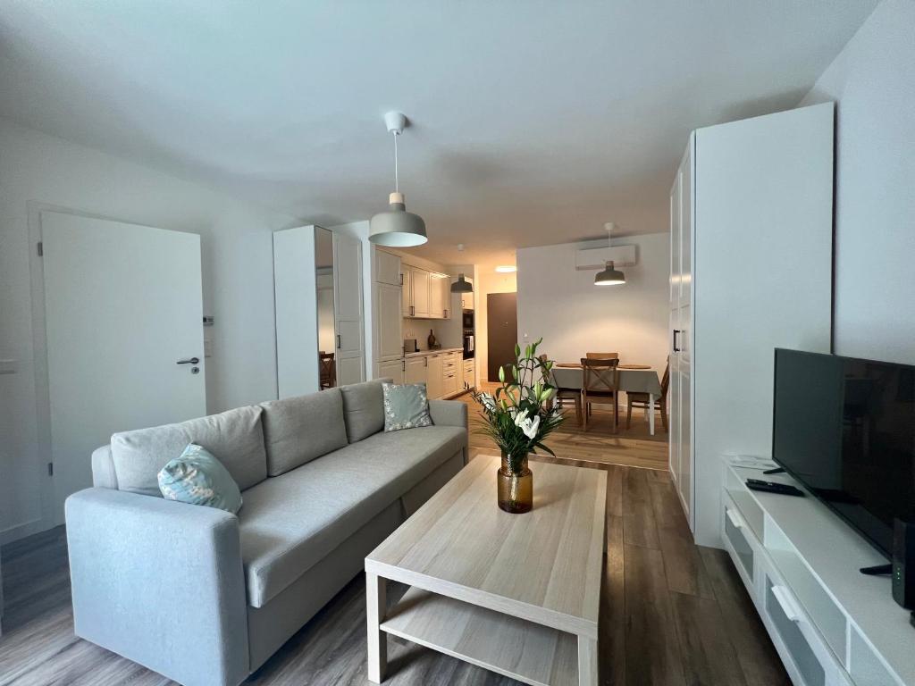 Uma sala de estar moderna com um sofá cinzento, mesa de centro de madeira, TV e cozinha aberta com zona de refeições ao fundo, no MT Suites ApartHotel.