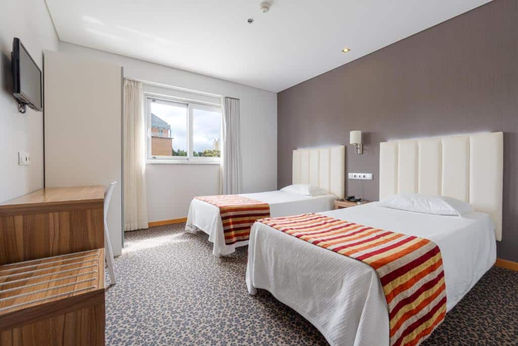 Quarto bem iluminado no Hotel Azinheira com duas camas individuais, colchas às riscas, uma secretária, TV na parede e uma janela com cortinados.