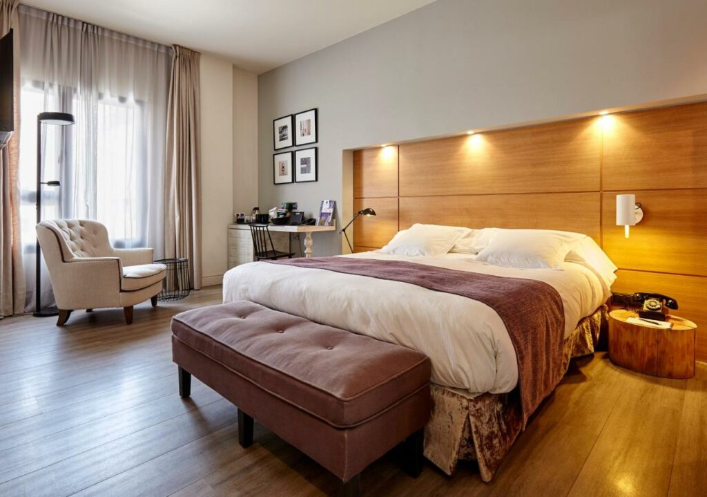 Quarto do Hotel Barcelona Catedral. A cama de casal encosta a uma cabeceira de madeira, com mesas de cabeceira de ambos os lados, um banco acolchoado aos pés e uma secretária junto a uma janela com cortinas bege.