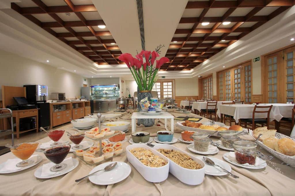 Uma mesa de bufete de pequeno-almoço com cereais, pães, doces e fruta numa sala de refeições bem iluminada com painéis de madeira no teto, servida no Consolata Hotel.