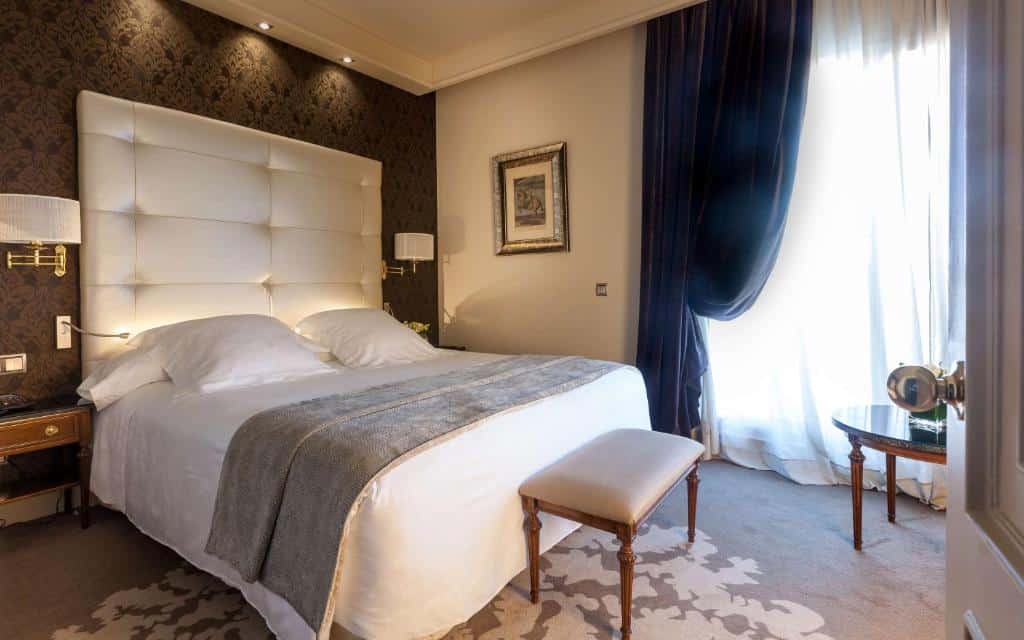 Quarto do Wellington Hotel & Spa Madrid, um dos melhores hotéis em Madrid. A cama de casal tem roupa de cama branca e cinzenta, cabeceira branca e mesinhas de cabeceira com candeeiro dos dois lados. Um banco está aos pés da cama. Na parede ao lado há uma janela coberta por cortinas brancas diáfanas e azuis, e um quadro ao lado. Há uma mesa redonda em frente à janela.