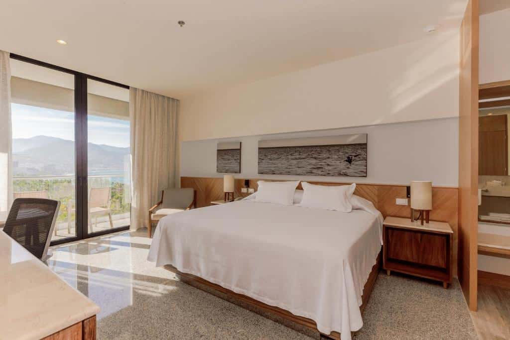 Quarto do Hotel Mio Vallarta com uma cama grande, duas mesas de cabeceira, uma cadeira, uma secretária e uma varanda com vista para as montanhas e a água.