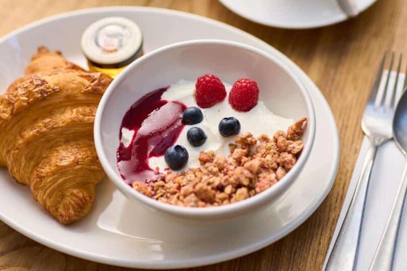 Refeição saudável e colorida no Trinity Townhouse Hotel, com croissant, iogurte com frutos vermelhos, granola e raspas de limão num prato branco sobre mesa de madeira.