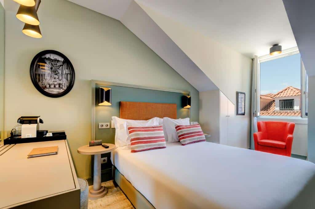 Quarto do Vincci Baixa com uma janela com cortinas e, por baixo, um cadeirão estofado, uma cama de casal, uma secretária do lado esquerdo com conjunto para preparar café e uma pufe