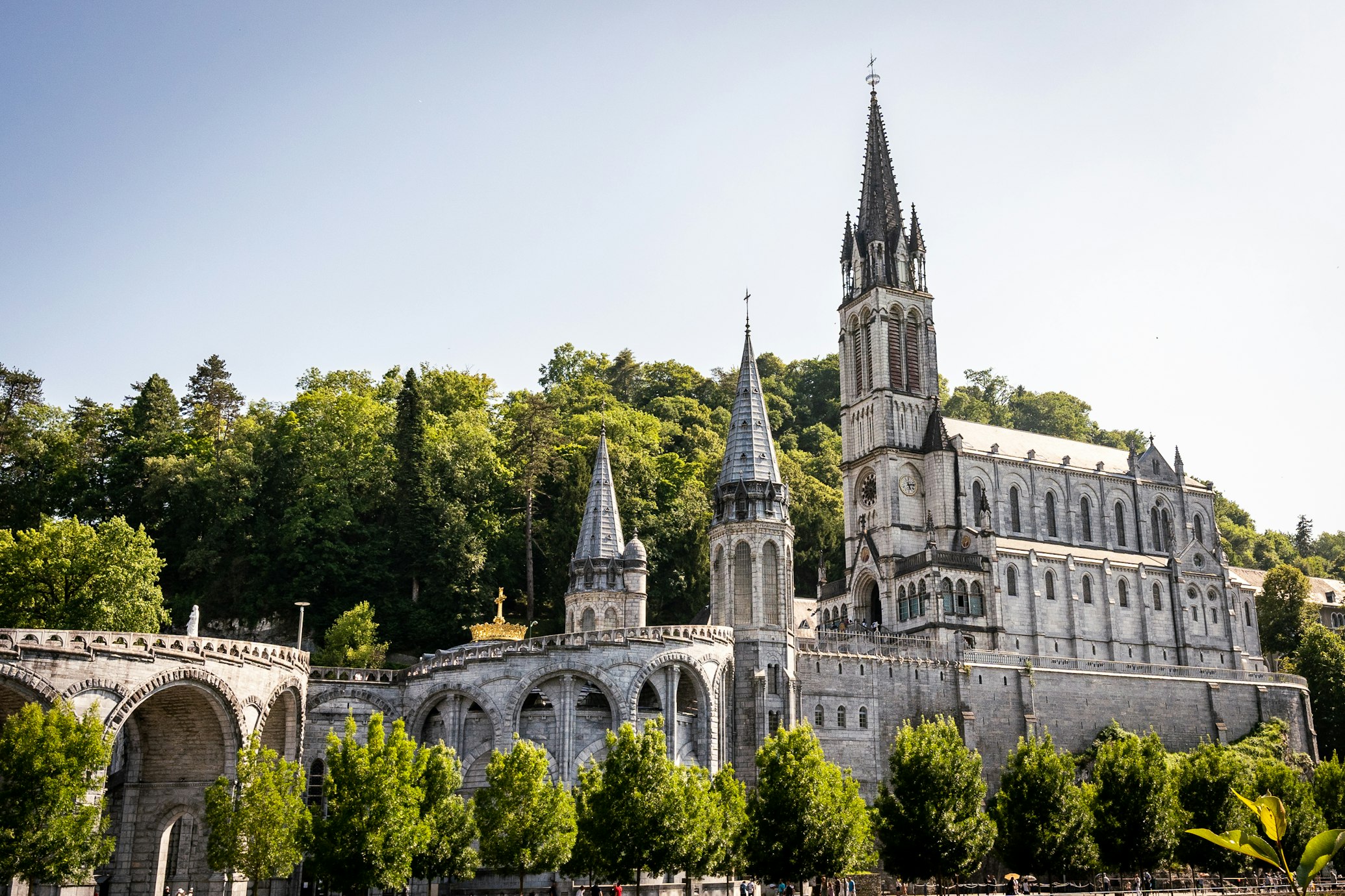 Grande igreja de pedra com várias torres e arcos, rodeada de árvores sob um céu claro, em Lourdes.