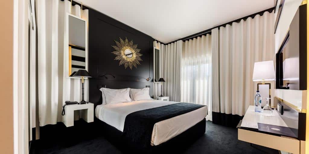 Quarto moderno no Aurea Fátima Hotel Congress & Spa com cama de casal, decoração a preto e branco, relógio de parede, candeeiros, secretária e grandes janelas com cortinados.
