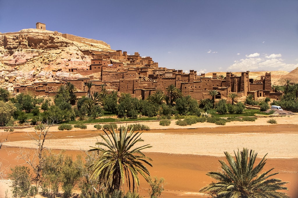 Antigos edifícios de Ksar Ait Benhaddou numa encosta, com palmeiras e um leito de rio seco em primeiro plano, sob um céu claro, em Marrocos.