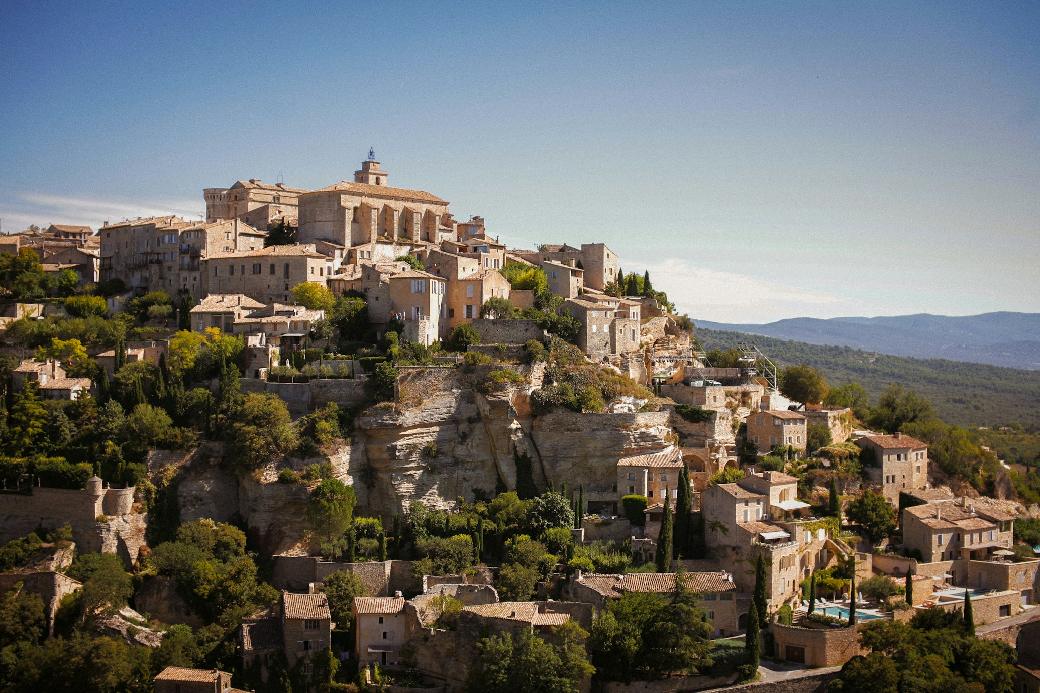 Aldeia no topo de uma colina, com edifícios de pedra e uma igreja proeminente, rodeada de árvores e com vista para um vale, em Gordes.