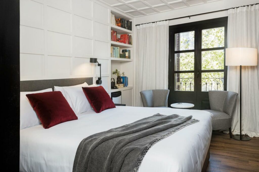 Quarto do Yurbban Ramblas Boutique Hotel, com cama de casal, candeeiros de ambos os lados, uma estante de livros e duas poltronas cinzentas junto a uma janela com cortinas brancas.