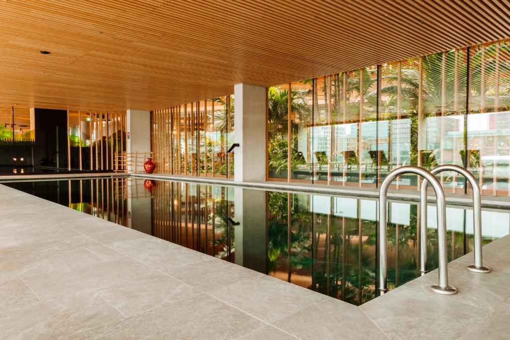 Piscina interior do Hotel Jakarta Amsterdam, com corrimãos de metal, pavimento em ladrilho, teto de madeira e janelas grandes voltadas para a vegetação.