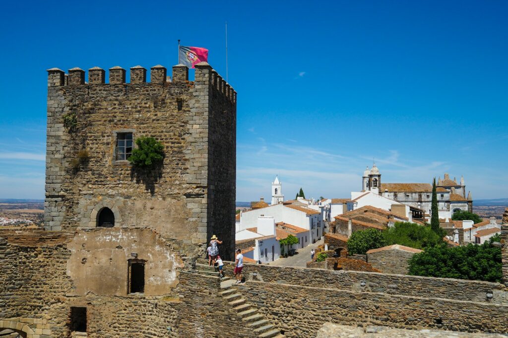 Castelo de Monsaraz, com uma bandeira de Portugal, em primeiro plano. A torre, de paredes de pedra escura, contrasta com as casas caiadas de branco e os telhados de telha ao fundo. Vêem-se pessoas a explorar o local, a dar vida à cena.