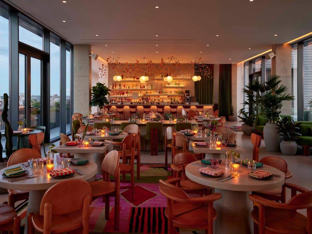 Restaurante do Penny Williamsburg com várias mesas redondas e cadeiras, cada uma decorada com velas. Ao fundo, um balcão de bar iluminado com prateleiras cheias de garrafas, grandes janelas de vidro, plantas e candeeiros suspensos.