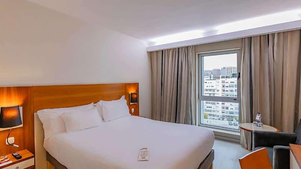 Quarto do Hotel Mercure Lisboa com cama grande, cabeceira de madeira e candeeiros de cabeceira, com janela com vista para a cidade.